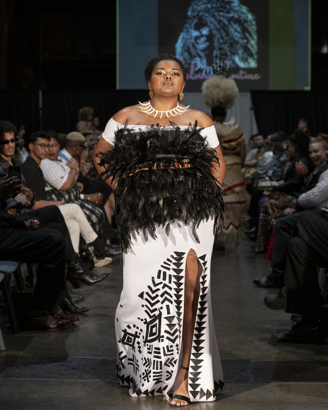 Model walking the Le Pasefika Runway in a white gown with black feather bodice and tapa patterns